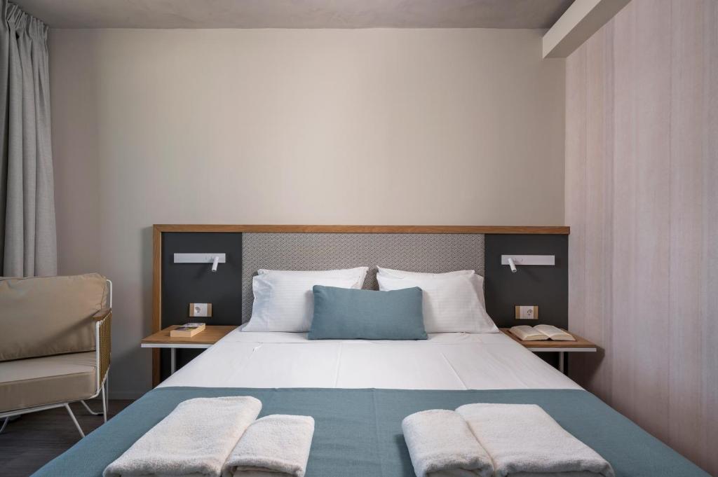 Mūsdienīga gulta ar baltām gultas veļām un zilu spilvenu, ērta krēsla stūrī un pie sienas piestiprinātiem gaismas avotiem. Svaiga un minimālistiska guļamistabas interjera dizaina estētika.