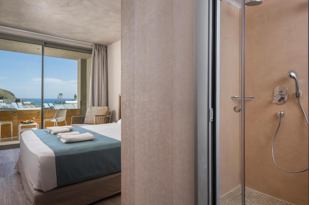 Mūsdienīgs viesnīcas numurs ar skatu uz jūru, kura interjerā ir ērta gulta un dušas kabīne. Numurā ir arī balkons ar atpūtas krēsliem.