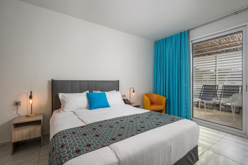 Mūsdienīga viesnīcas istaba ar gaišām krāsām, ērtu gultu un dīvānu, kas vērsts uz balkonu. Logā redzams ārējais terases laukums ar sēdvietām.