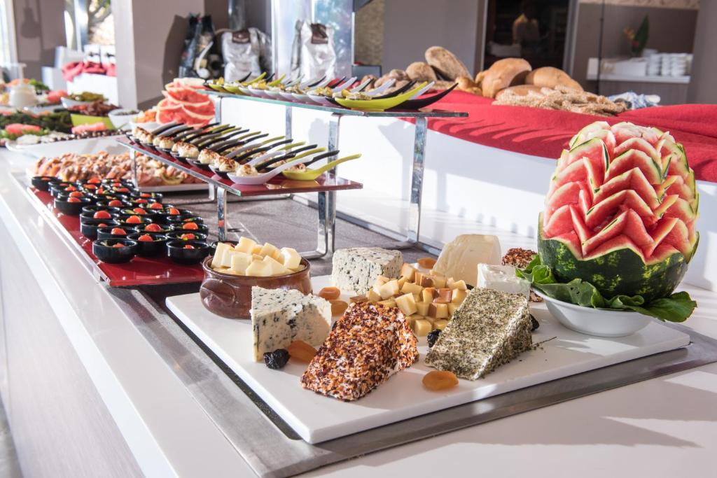 Bagātīgs uzkodu galds ar dažādām sieru šķirnēm, svaigiem dārzeņiem un augļiem, tostarp dekoratīvi sagriezta arbūza. Galdā ir arī maizītes un citas gardēžu uzkodas.