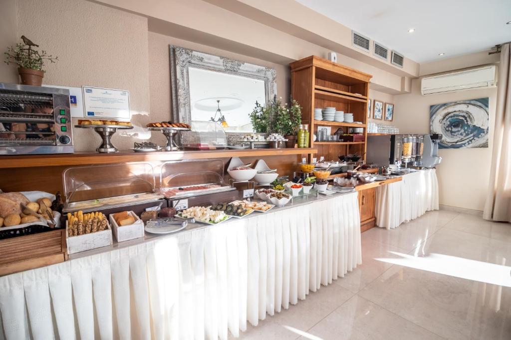 Bagātīgs brokastu bufete ar dažādiem ēdieniem, tostarp maizi, augļiem un desertiem, sakārtota pie elegantas koka plaukta. Gaisma plūst caur telpu, radot siltu un viesmīlīgu atmosfēru.