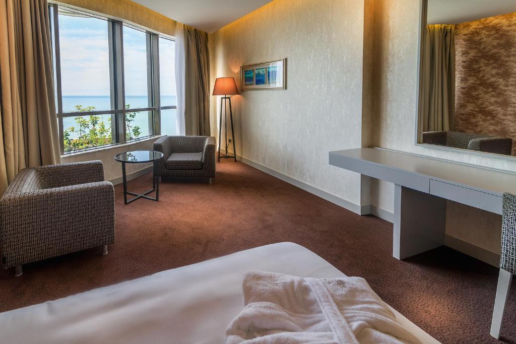 Grezns viesnīcas numurs ar skatu uz jūru, kurā ir divi ērti krēsli, kafijas galdiņš un liels spogulis. Telpa ir gaišā tonī ar mūsdienīgu interjeru un dabiskiem apgaismojumiem.