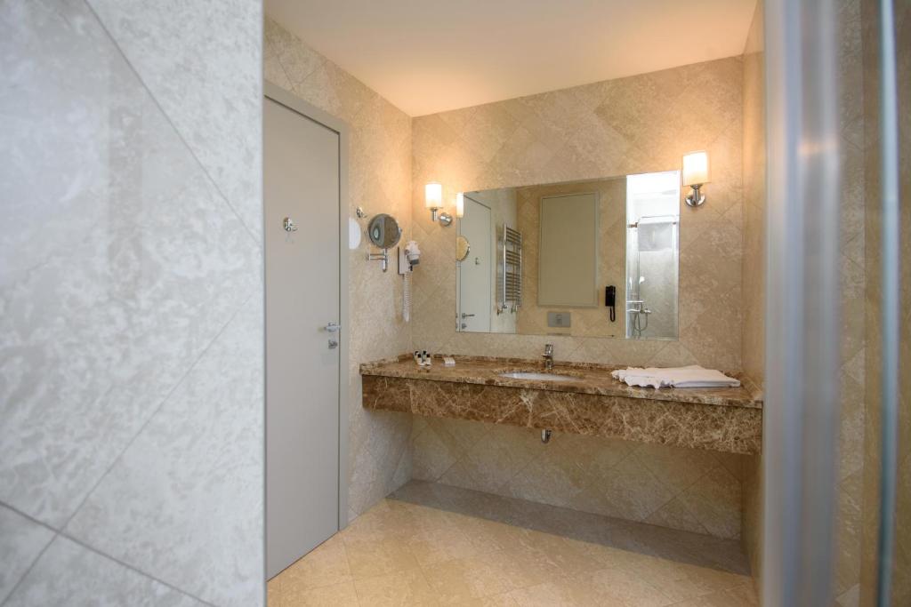 Mūsdienīgs vannas istabas interjers ar marmora izlietni un spoguli, kas izgaismots ar siltām gaismām. Redzamas arī sienas flīzes un durvis, kas piešķir telpai elegantu izskatu.