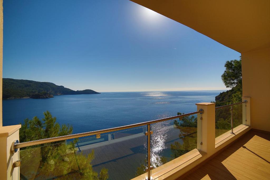 Skats no balkona uz mierīgu jūras ainavu ar saules gaismu, kas atspoguļojas ūdenī. Fons veido kalnu kontūras un zilo debesu loku.