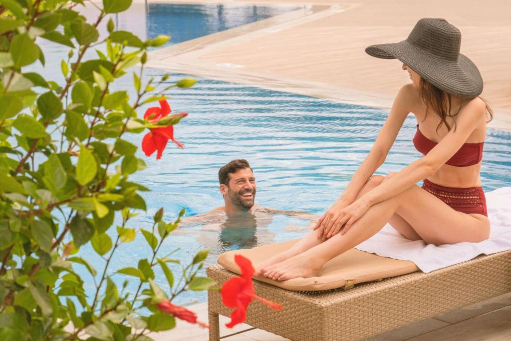 Atpūtas brīdis pie baseina, kur sieviete sēž uz sauļošanās krēsla, bet vīrietis smejas un atpūšas ūdenī. Apkārt ziedi un zaļumi rada relaksējošu atmosfēru.