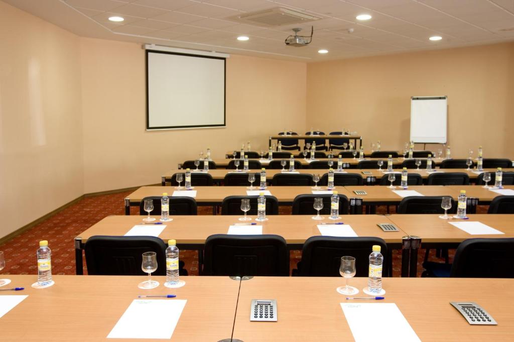 Konferenču telpa ar galdiem, krēsliem un projektoru, gatava sanāksmēm un prezentācijām. Uz galdiem ir ūdens pudeles, glāzes un piezīmju lapas.