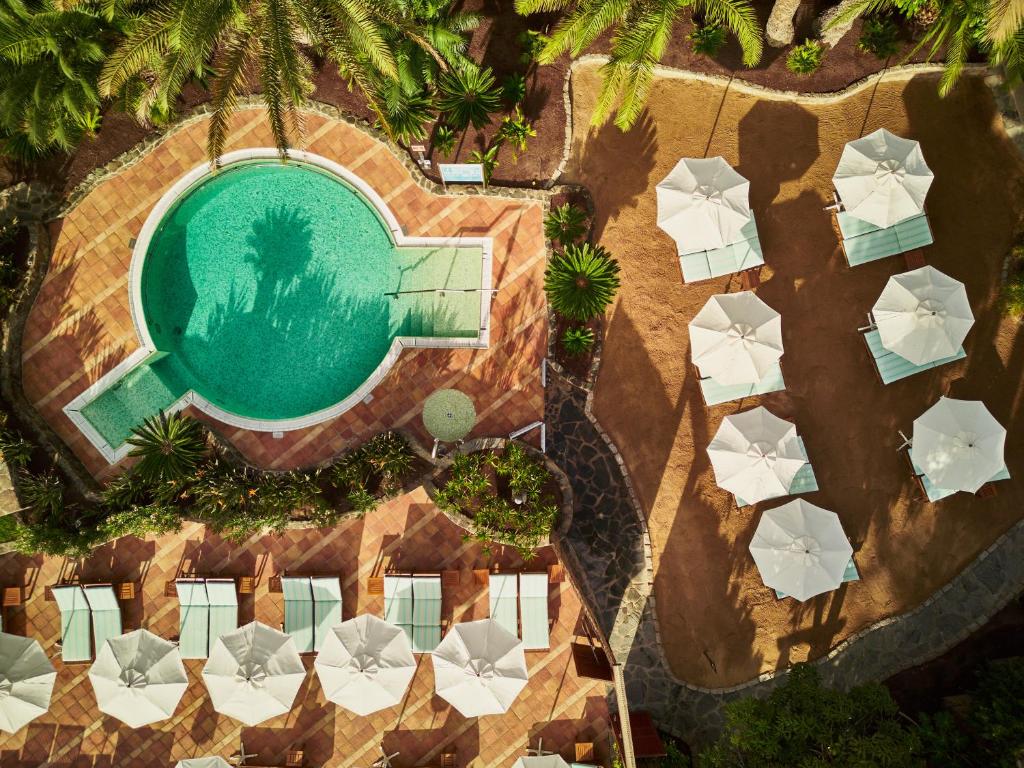 Peldbaseins ar palmām un sauļošanās krēsliem, kas izvietoti ap to, radot relaksējošu atmosfēru. Ūdens atspoguļo saules gaismu, kamēr blakus ir saulessargi smiltīs.