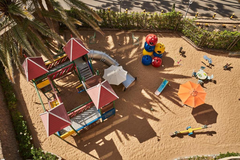 Bērnu spēļu laukums ar krāsainām struktūrām, slidkalniņu un smilšu segumu, ko ieskauj palmas. Redzami rotaļu elementi, piemēram, šūpoles un galdiņu komplekts.