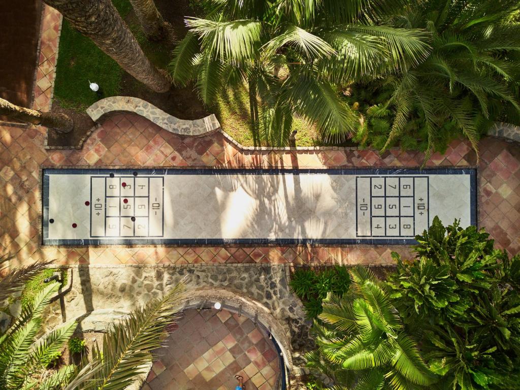 Šī ir gaisa skats uz šadžu laukumu, kas atrodas dārzā, ko ieskauj tropiskas augu lapas. Laukums ir izklāts ar flīzēm un atzīmēts ar skaitļiem un līnijām.