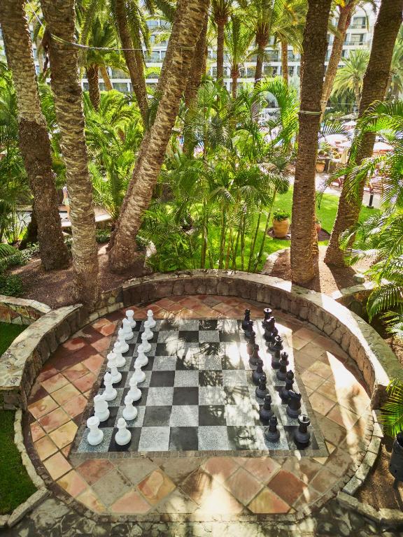 Āra šaha laukums ar lielām melnām un baltām figūrām, ko ieskauj tropiskas palmas un sulīgas augu ainavas. Ideāla vieta relaksācijai un spēlēm.