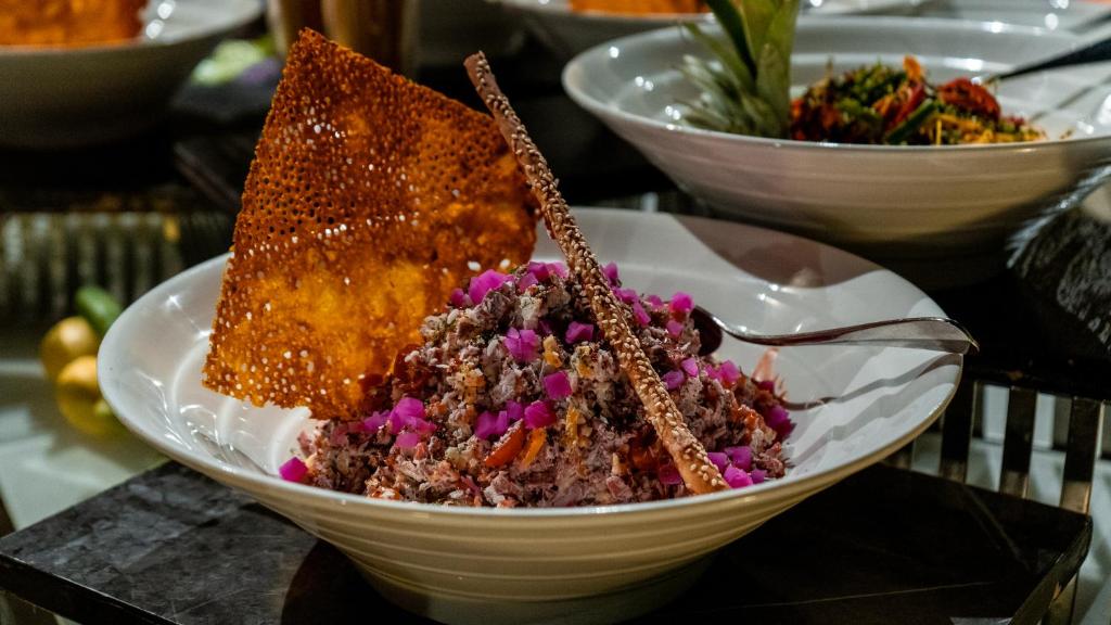 Grezna plāksne ar gaļas ēdienu, dekorēta ar rozā ziedlapiņām un kraukšķīgu čipsu. Uz galda redzami arī citi ēdieni, radot kulinārijas svētku noskaņu.