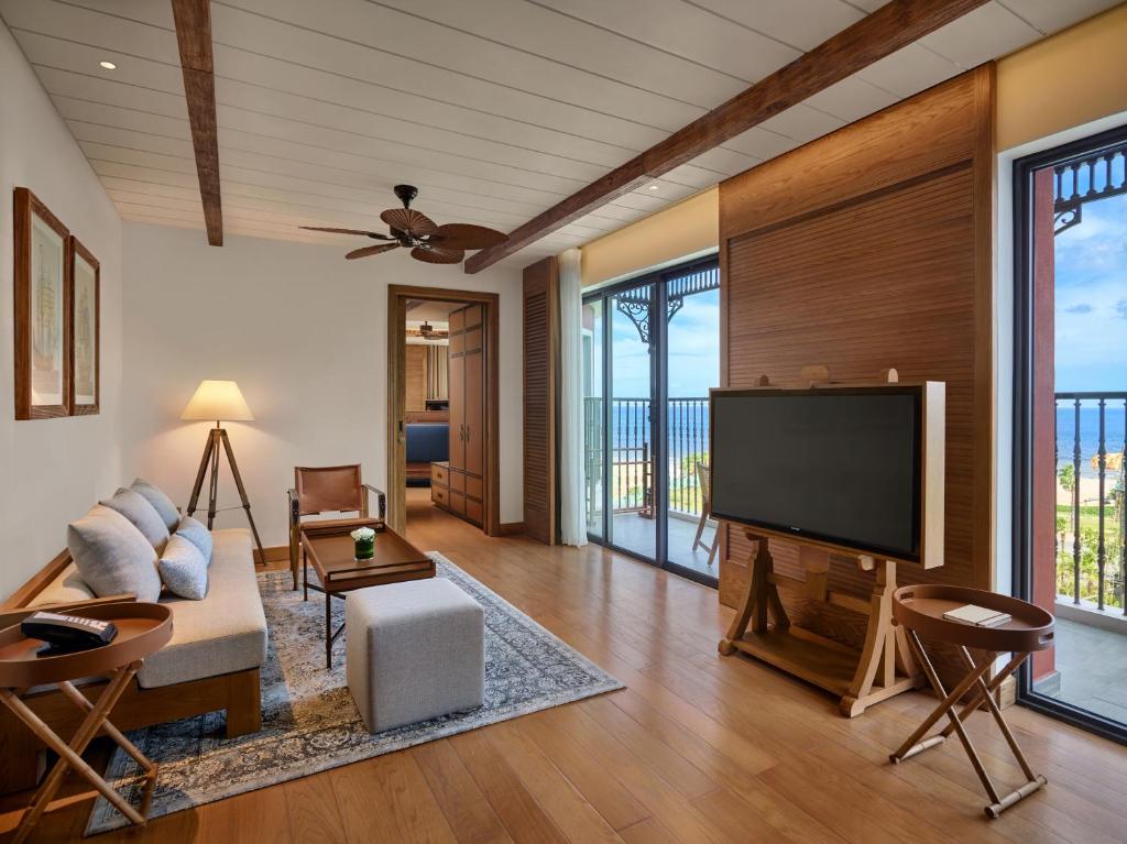 Mūsdienīgs viesistabas interjers ar plašiem logiem, kas sniedz skatu uz jūru. Telpā ir komfortabla dīvāna zona, koka elementi un televizors uz statīva.