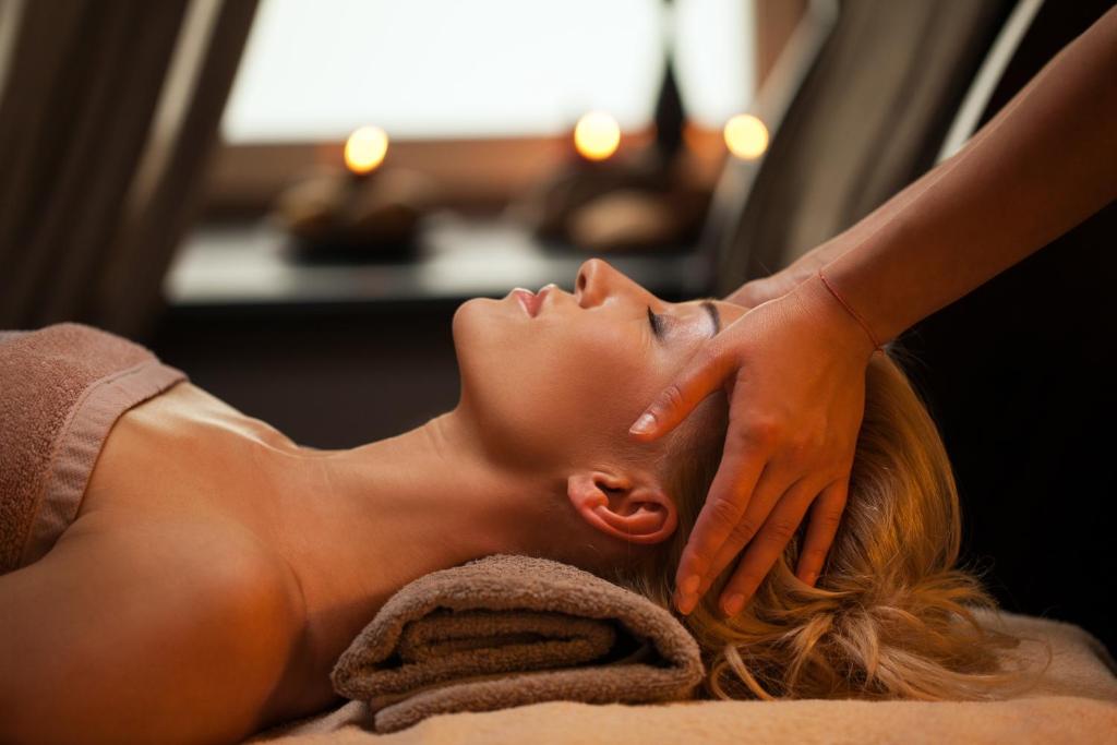 Relaksējoša masāža spa centrā, kur sieviete saņem galvas masāžu, kamēr atpūšas uz dvieļiem. Apkārt ir svecītes, radot mierīgu atmosfēru.