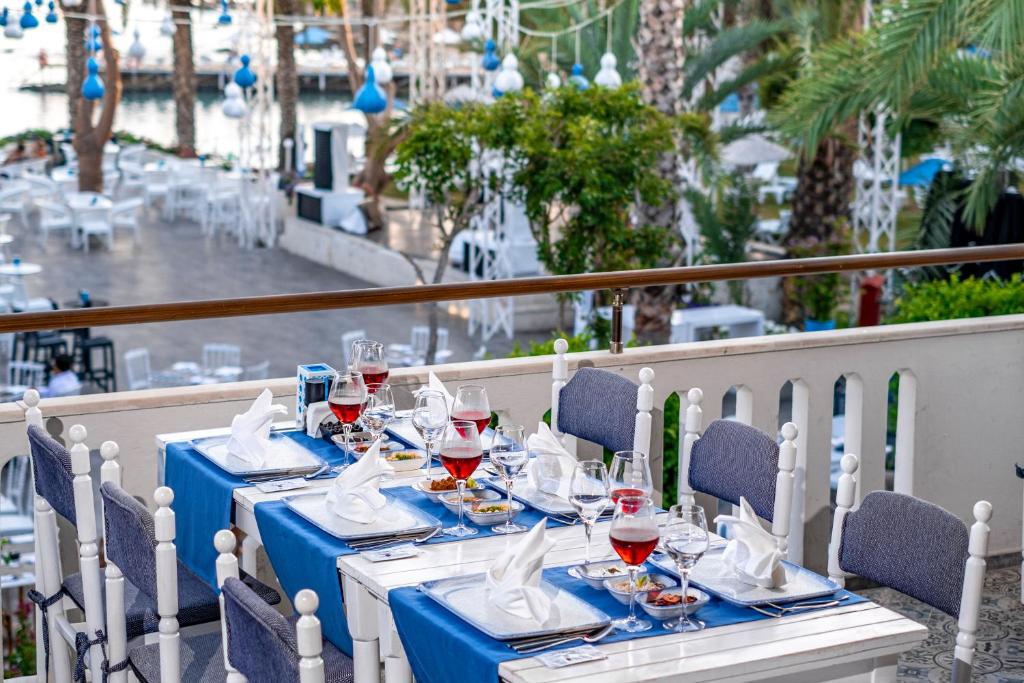 Elegants restorāna galds ar sarkanvīna glāzēm un dekoratīvām salvetēm, aiz kura redzami palmām un zilo gaismu rotāti ārējie sēdvietas. Ideāla atmosfēra relaksējošai vakariņai pie ūdens.