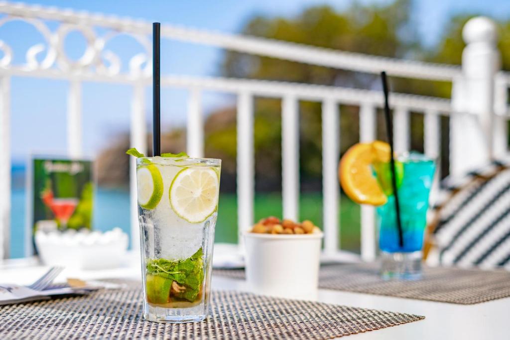 Atspirdzinoši kokteiļi, tostarp mojito un zils kokteilis, tiek pasniegti pie galda ar riekstiem un skaistu ainavu fona.