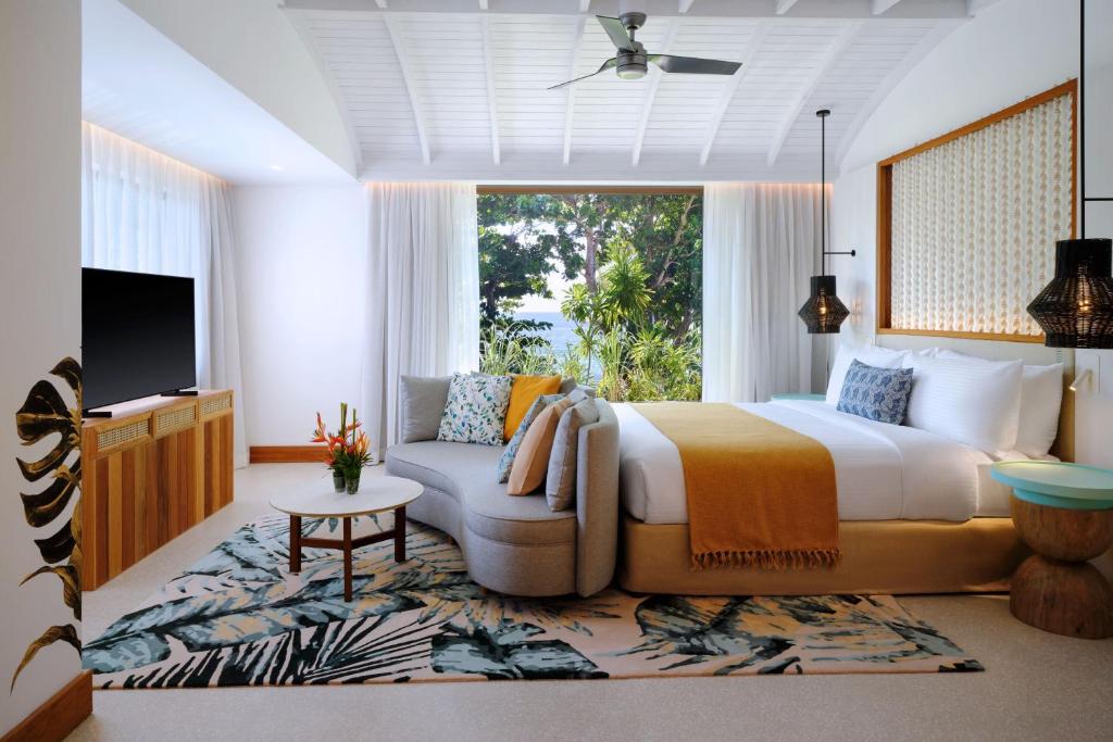 Mūsdienīgs viesnīcas numurs ar gultu, dīvānu un televizoru, kas atrodas pie liela loga ar skatu uz dārzu. Interjers ir gaišs un elegant, ar augiem un dekoratīviem elementiem.