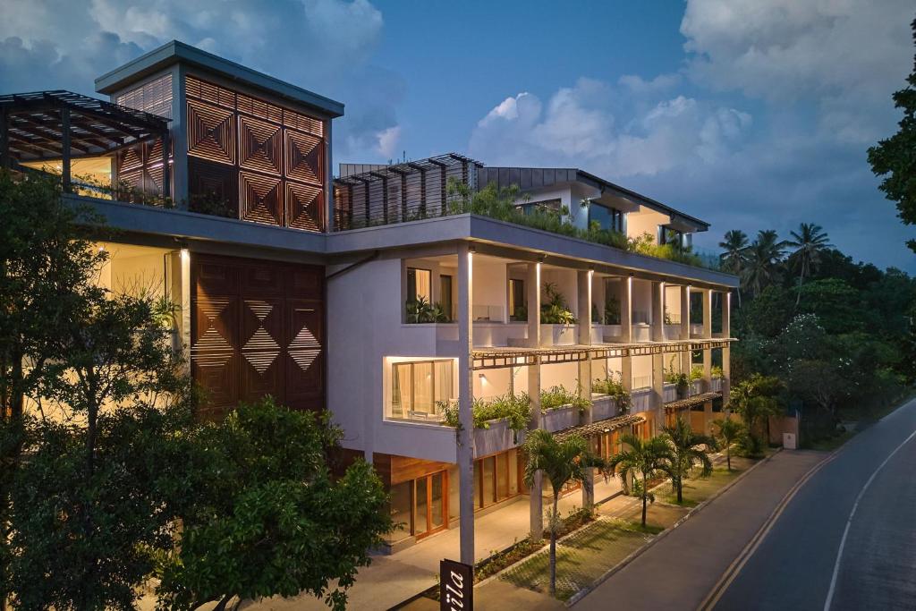 Moderns viesnīcas kompleks, kas ieskauts zaļumā, ar arhitektūras detaļām, kas izceļas vakarā. Apgaismojums akcentē eleganto dizainu un viesmīlīgu atmosfēru.