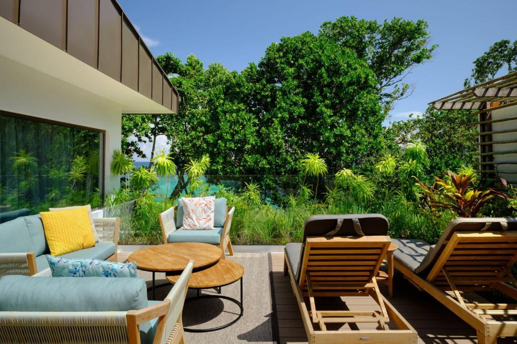 Mūsdienīgs terases interjers ar ērtiem dīvāniem, apaļu galdu un sauļošanās krēsliem, apkārt zaļojošiem augiem un skatu uz ūdeni.