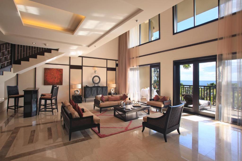 Mūsdienīgs viesistabas interjers ar augstiem logiem un skatu uz jūru. Telpa ir eleganti iekārtota ar mīkstajiem dīvāniem, galdu un barstūri.