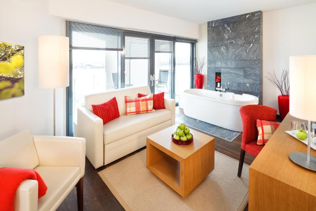 Mūsdienīgs viesnīcas numurs ar elegantu interjeru, baltu dīvānu un sarkanām akcentu krēsliem. Telpā atrodas arī brīvstāvoša vanna un koka galdiņš ar zaļām āboliem.