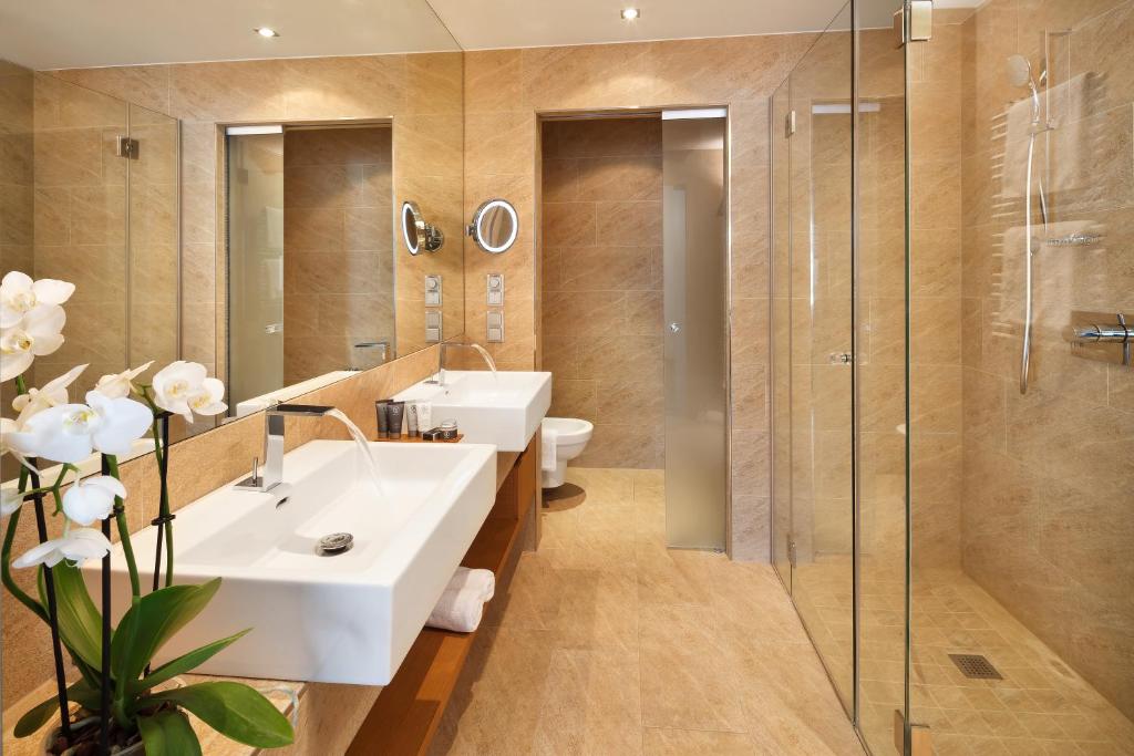 Mūsdienīgs vannas istabas interjers ar diviem izlietnēm, stikla dušu un dabiskām orhidejām uz letes. Siltas krāsas un eleganta dizaina elementi rada relaksējošu atmosfēru.