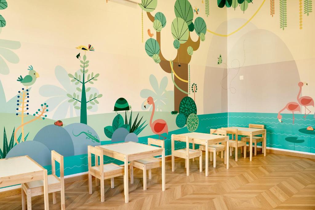 Bērnu istaba ar krāsainu sienu gleznojumu, kas attēlo tropisku dabu un dzīvniekus, un koka galdiem un krēsliem. Ideāla vide radošām aktivitātēm.