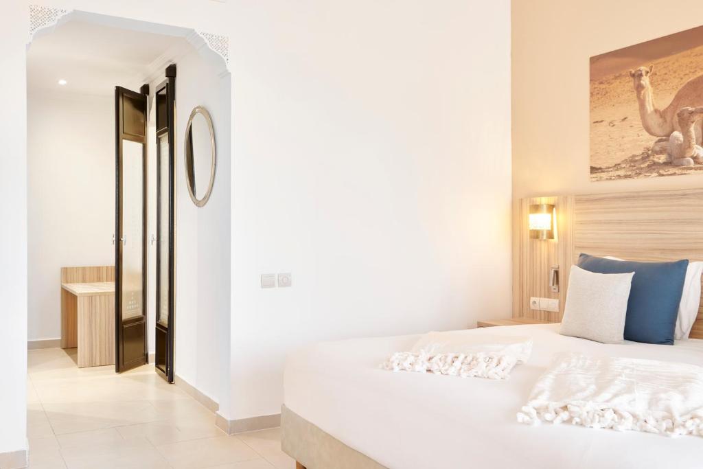 Mūsdienīga guļamistaba ar gaisīgu interjeru un piekļuvi vannas istabai, kurā ir koka mēbeles un liels spogulis. Sienā redzama gleznainā kamieļa fotogrāfija.