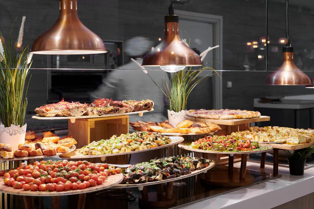 Bufete ar dažādiem ēdieniem, tostarp dārzeņiem, maizēm un uzkodām, apgaismota ar stilīgām lampām. Šī attēla atmosfēra ir viesmīlīga un pievilcīga.