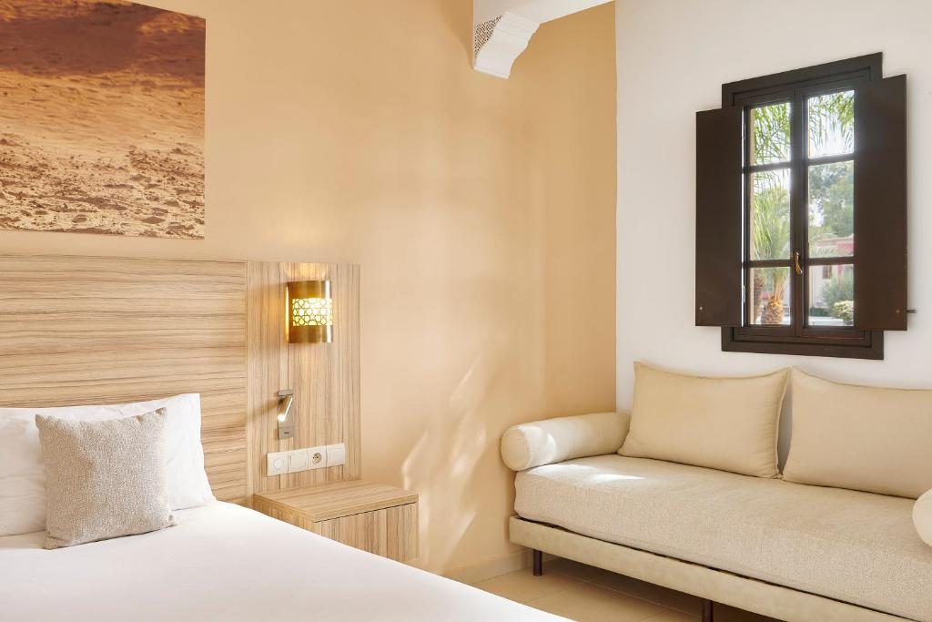 Moderns viesnīcas numurs ar gultu, sēdvietu un logu, kas piedāvā skatu uz dārzu. Sienas ir krāsotas smilšu krāsā, radot siltu un viesmīlīgu atmosfēru.