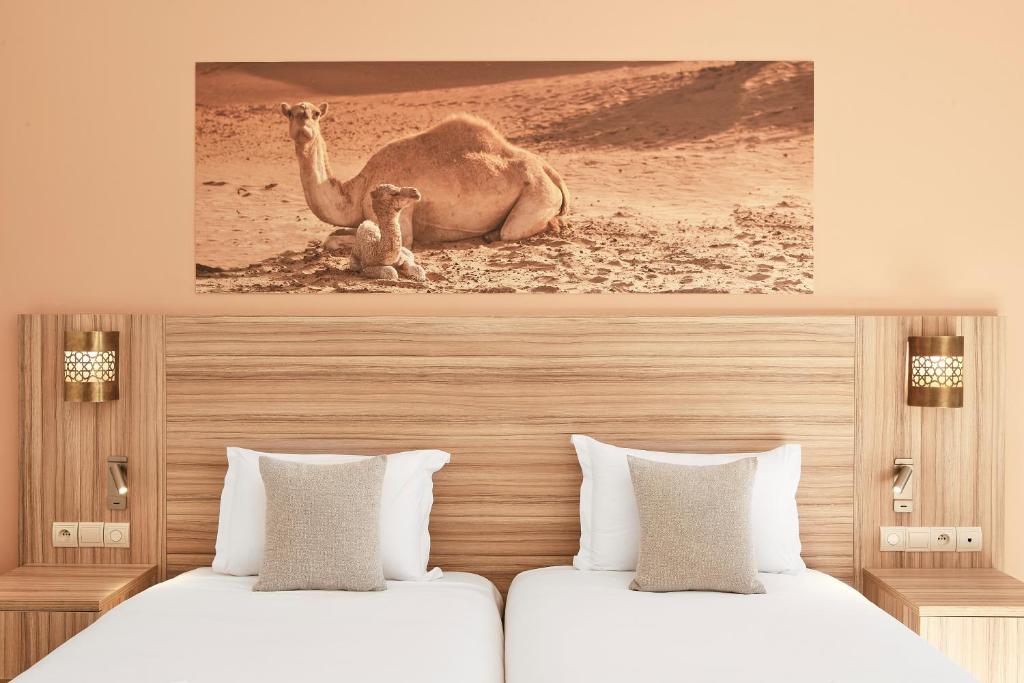 Mūsdienīga viesnīcas numura interjers ar divām gultām un kameļu attēlu uz sienas. Siltas krāsas un eleganti aksesuāri rada patīkamu atmosfēru.