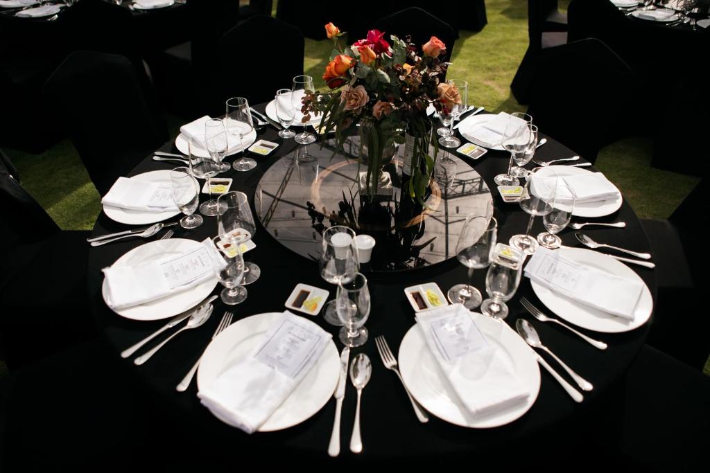 Eleganti izklāta svinību galda kompozīcija ar melnu galdautu, baltiem šķīvjiem un ziedu kompozīciju centrā. Ap galdu novietotas glāzes un galda piederumi, radot svinīgu atmosfēru.