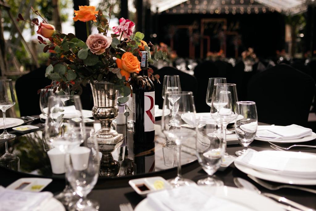 Elegants galda dekors ar ziedu kompozīciju un vīna pudeli, kas radījis svinīgu atmosfēru. Galdu klāj melni galdauti un izsmalcināta galda piederumi.