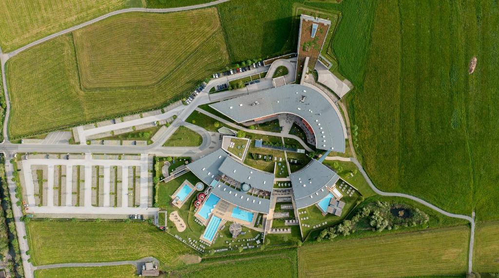 Gaisa skats uz modernu viesnīcu kompleksu, kas iekārtots zaļajā lauku vidē, ar peldbaseiniem un plašām stāvvietām. Apkārtne ir gleznaina un labi koptā.