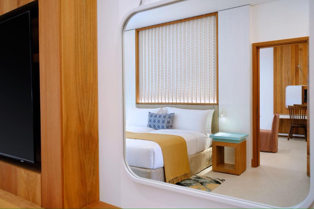 Mūsdienīgs guļamistabas interjers ar koka akcentiem, ērku gultu un rakstāmgaldu, redzams spogulī. Siltas krāsas un minimālisma stils rada relaksējošu atmosfēru.