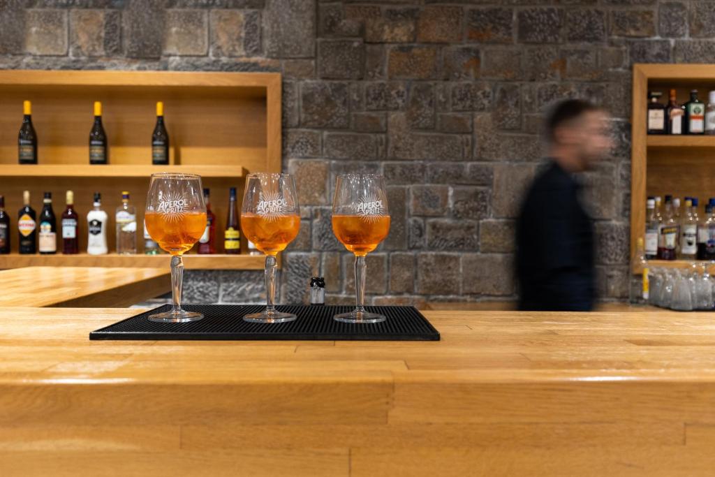 Trīs Aperol Spritz glāzes stāv uz bāra, fonā redzama neskaidra persona un dažādas alkohola pudeles.