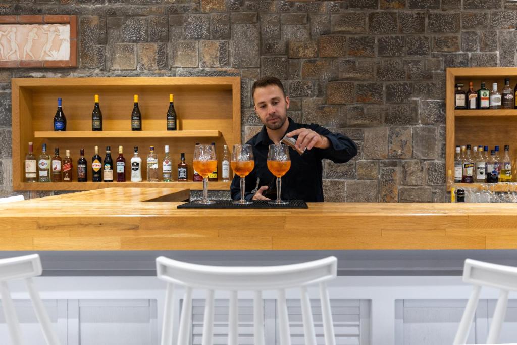 Bāra darbinieks gatavo kokteiļus, stāvot pie moderna bāra ar koka letēm un akmens sienu. Fona plauktā redzami dažādi alkoholiskie dzērieni.