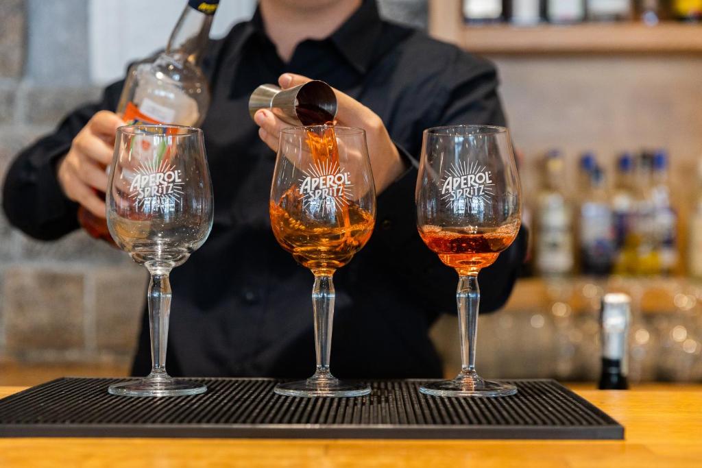 Barmenis ielej Aperol Spritz kokteili trīs glāzēs, radot dzirkstošu un dzīvīgu atmosfēru. Fons satur dažādas alkoholiskās dziras, kas piešķir bāra noskaņu.