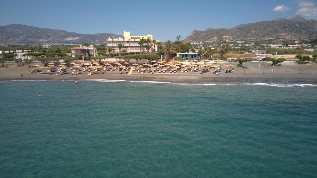 Pludmale ar sauļošanās krēsliem un lietussargiem, fons ar kalniem un viesnīcu. Skaidrs zils ūdens un saulains laiks.