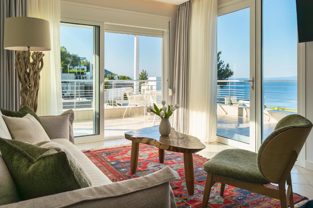 Mūsdienīgs viesistabas interjers ar skatu uz jūru, kurā ir ērta dīvāna, koka galds un ziedu kompozīcija. Gaismas plūst cauri lielajiem logiem, radot svaigu un atvērtu atmosfēru.