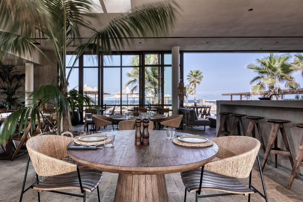 Mūsdienīgs restorāns ar skatu uz pludmali, kurā atrodas eleganta koka galda un ērti krēsli. Interjeru rotā zaļie augi un lieli logi, kas ļauj ieplūst dabiskajai gaismai.