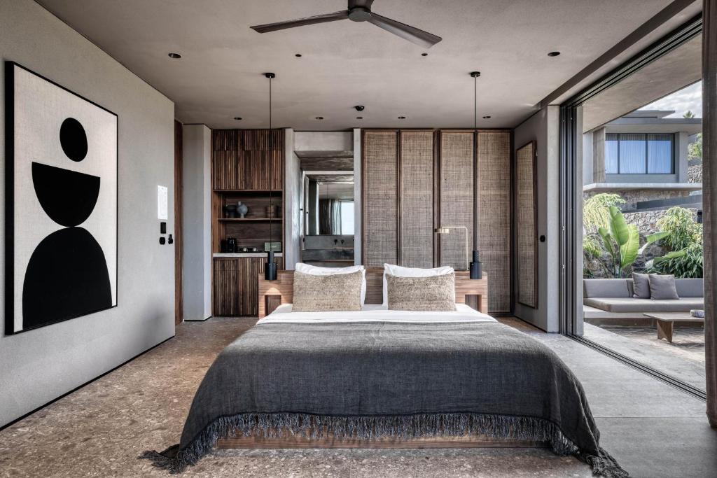 Mūsdienīga guļamistaba ar minimālisma dizainu, kurā ir liels gultas matracis, dekoratīvi spilveni un mākslas darbs uz sienas. Atvērta telpa ar skatienu uz āru un dabas elementiem.