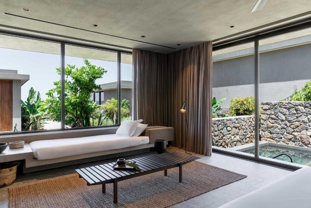 Mūsdienīgs viesnīcas numurs ar lieliem logiem un skatu uz dārzu, kur atrodas akmens mūris un zaļumi. Interjers ir elegantā stilā ar minimālistisku mēbelējumu un dabīgiem materiāliem.