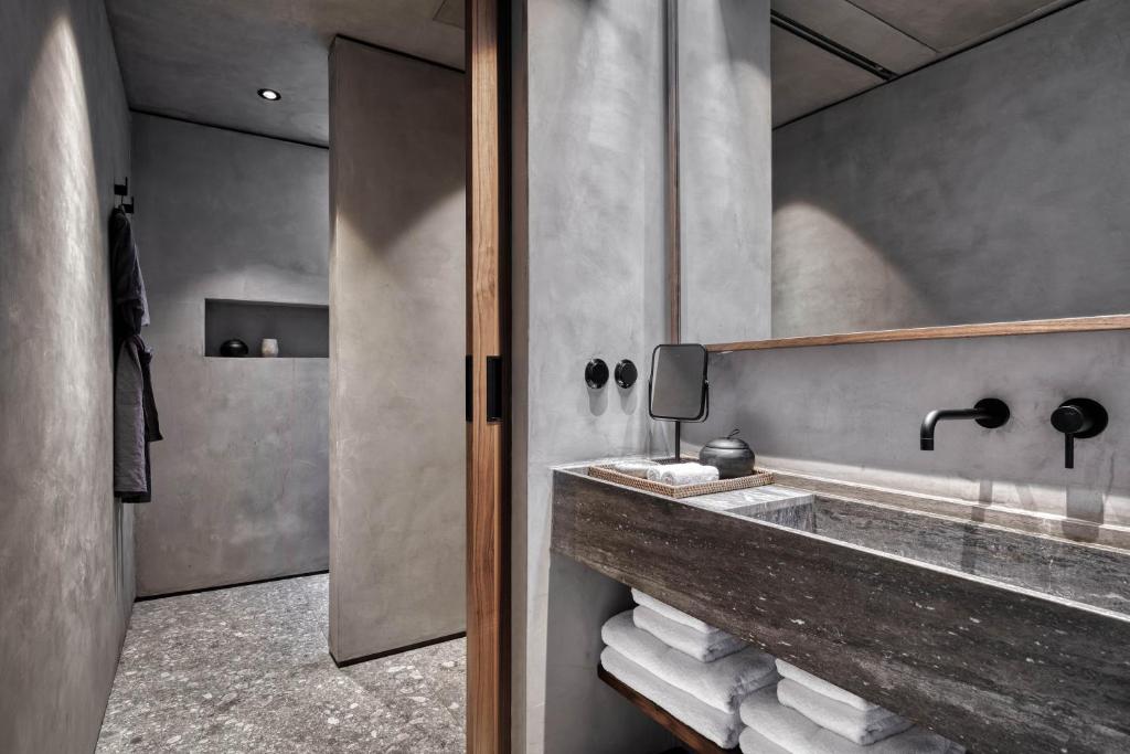 Mūsdienīgs vannas istabas interjers ar betona sienām un elegantu akmens izlietni, papildināts ar melnām ūdens krāniem un koka elementiem. Gaisma ir maiga, radot relaksējošu atmosfēru.