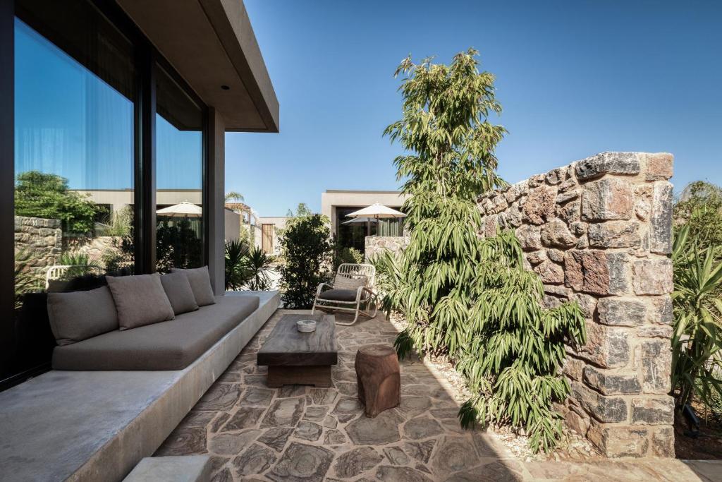 Mūsdienīgs terases stūris ar akmens sienu, ērtu dīvānu un dabas elementiem, kas rada relaksējošu atmosfēru. Papildina zaļumi un saulainais debesis.