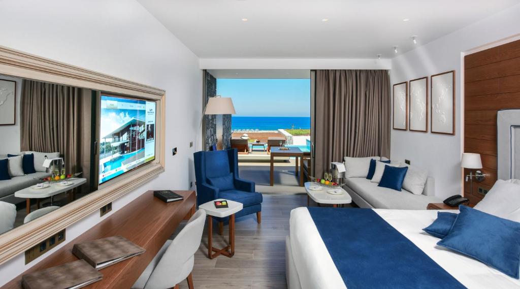 Mūsdienīga viesnīcas istaba ar skatu uz jūru, gaišiem toņiem un stilīgu mēbelējumu. Telpā ir gulta, dīvāns un televizors, kā arī balkons ar piekļuvi baseinam.