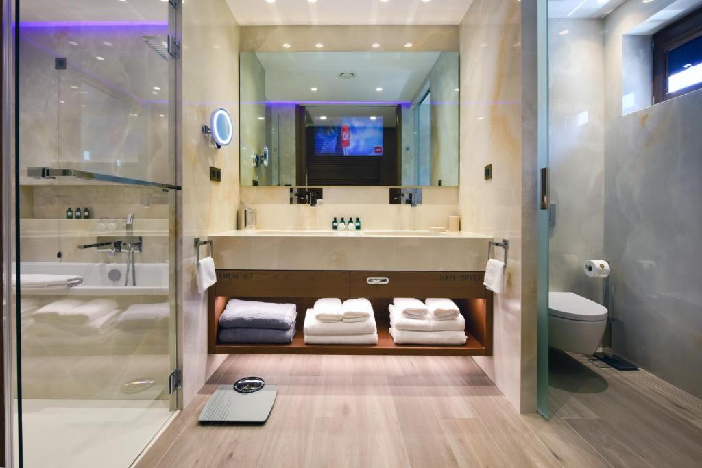 Mūsdienīga vannas istaba ar stikla dušas kabīni, elegantu izskatu un mīkstiem dvieļiem. Uz galda redzami grezni higiēnas piederumi un spogulis ar apgaismojumu.