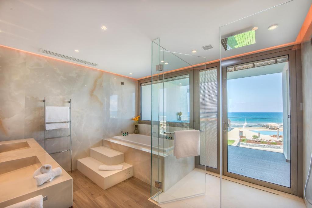Mūsdienīgs vannas istabas dizains ar stikla sienām un skatu uz jūru. Telpā ir vannas un mūsdienīgas apdares detaļas, radot relaksējošu atmosfēru.