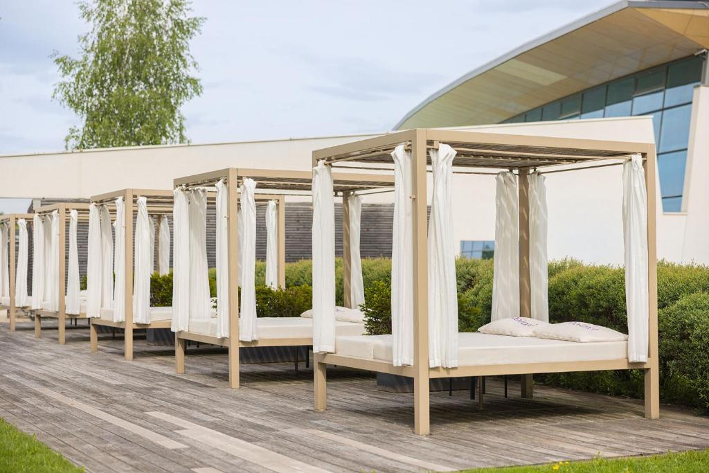 Atpūtas gultas ar baltām aizkarām, izvietotas uz koka klāja pie modernas ēkas, apkārt zaļojošas krūmu grupas.