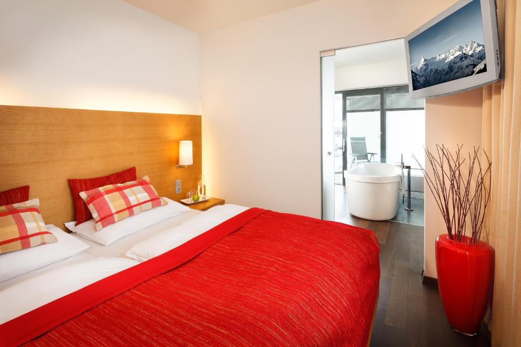 Mūsdienīga guļamistaba ar sarkanu gultas veļu un ērtiem spilveniem, kas piedāvā skatu uz balkonu un vannu. Interjers ir gaišs ar koka akcentiem.