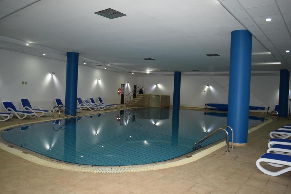 Iekštelpu baseins ar zilu flīzēm un baltām sauļošanās krēsliem, apgaismojums radīs relaksējošu atmosfēru. Ideāla vieta atpūtai un izklaidei.